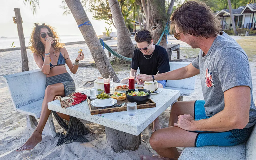 Im Urlaub schlemmen – Ja, aber bitte gesund | Drei Personen sitzen am Tisch am Strand und essen gesund im Schatten der Palmen